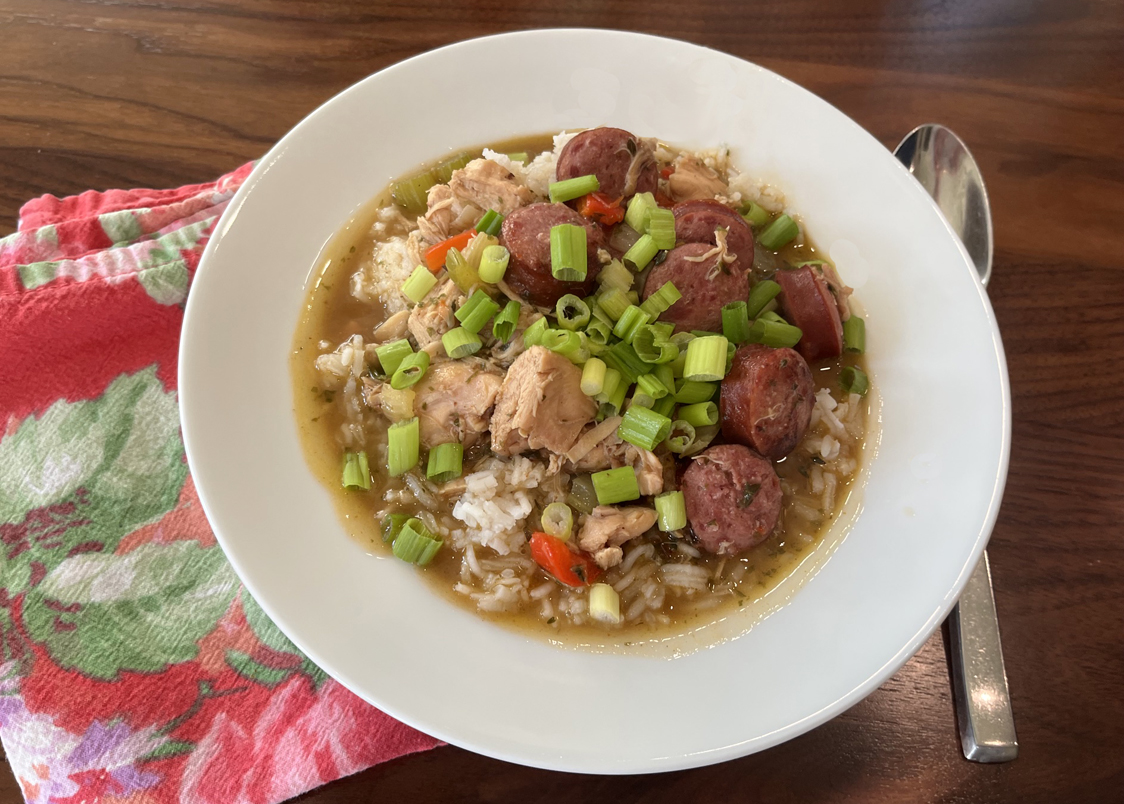 Image shows a white bowl with a pink floral napkin and a spoon beside it. In the bowl is gumbo, with cihcken, sausage and green onions.