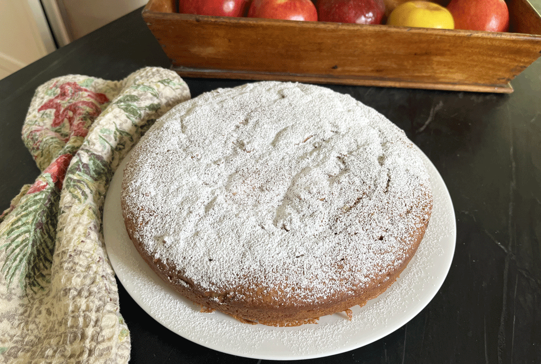 A round, flat cake dusted with powdered sugar on a white plate, with a wooden container of apples nearby.