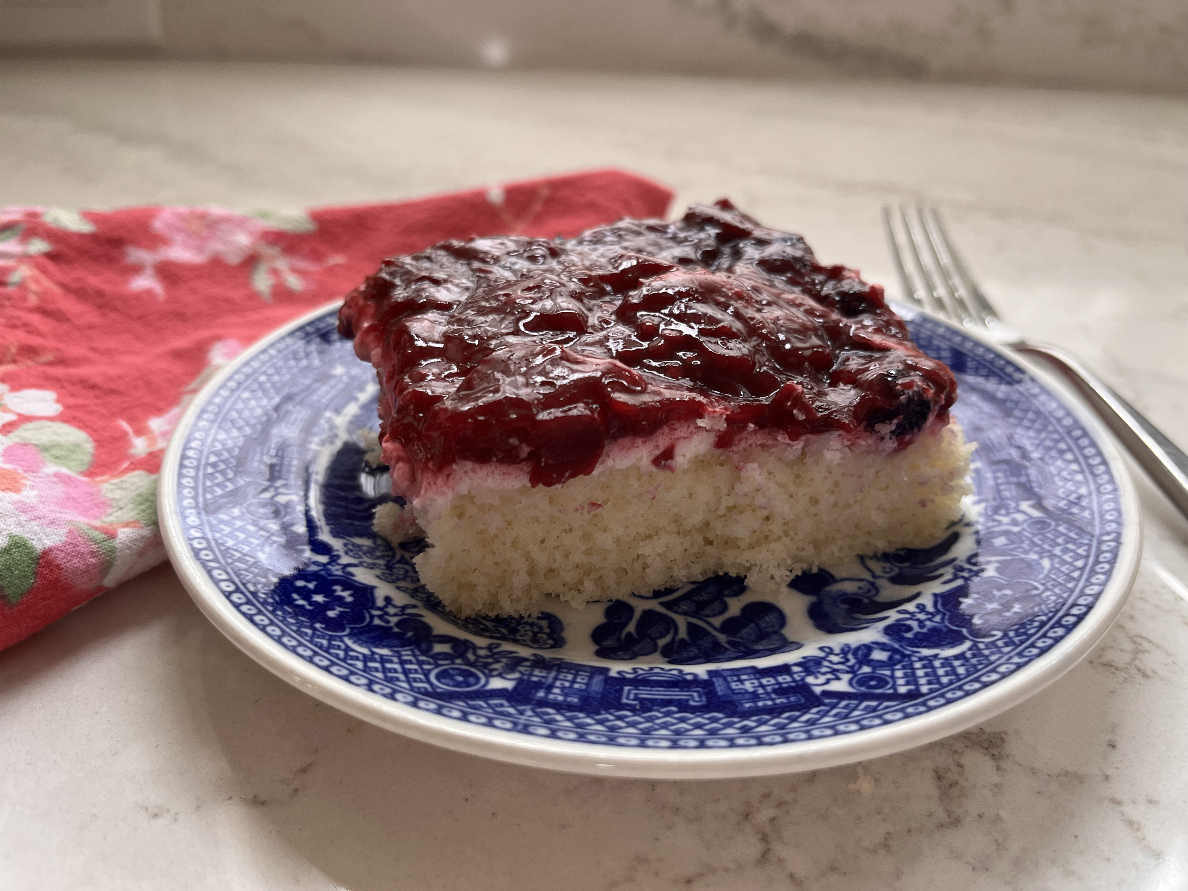 photo of a white cake with a cream layer and a berry topping, served on a blue plate with a pink napkin and a fork