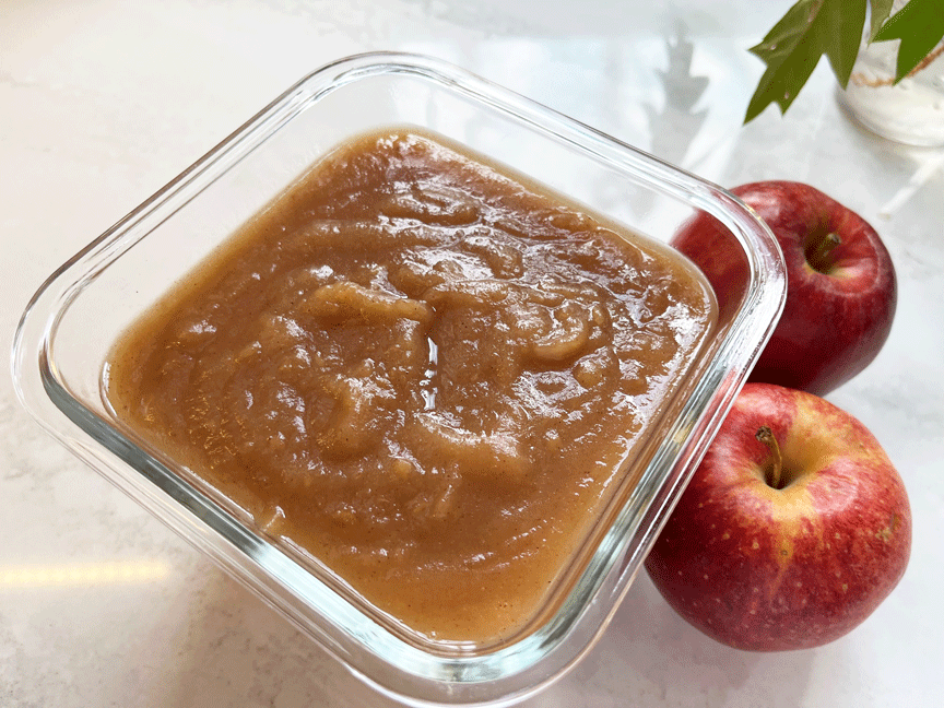 Cinnamon applesauce in a clear glass storage container with 2 apples