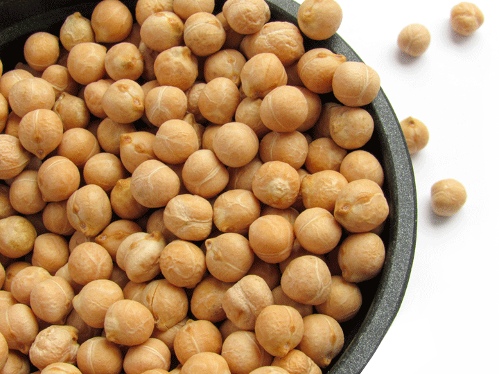 Photos shows a bowl of dried garbanzo beans in a dark ceramic bowl, with a few spilled out on the side.