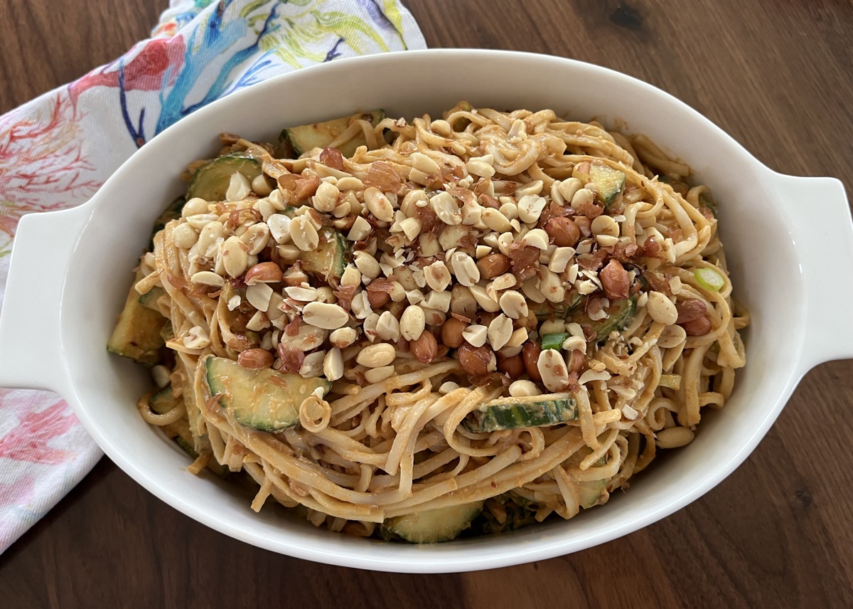 image of a spaghetti, cucumbers and peanuts in a white oval bowl with a colorful cloth nearby and on a wood background.