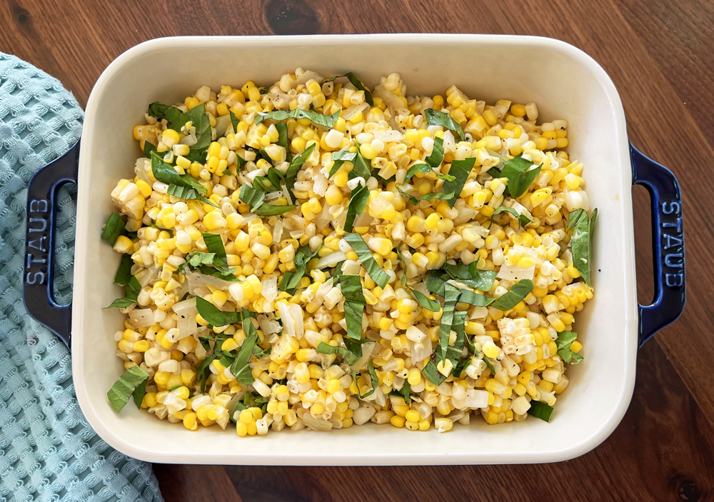 Image of a blue dish on a wooden table with a seafoam waffle hand towel. In the dish is a salad with corn, onions and basil leaves.