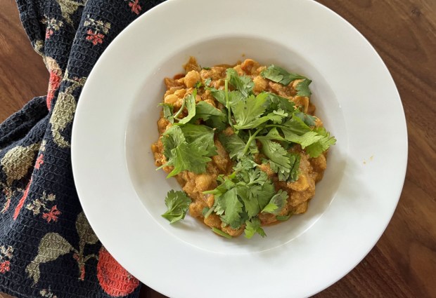 Garbanzo bean curry with chopped cilantro on top, in a white bowl on a wood table with a floral cloth beside the bowl.