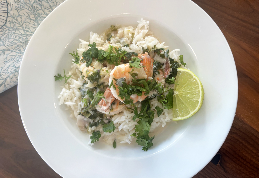 A white bowl on a wooden table, with rice, shrimp, cilantro and a wedge of lime on the side. 
