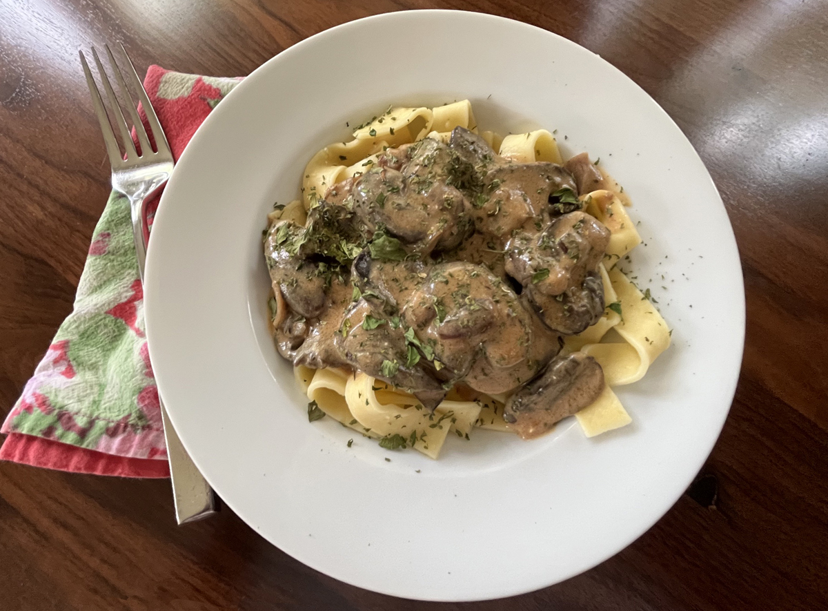 Photo shows a white bowl of wide noodles topped with a gravy and mushrooms.  A pink floral napkin is beside the bowl with a fork on a wooden table.